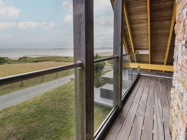 A balcony with wooden floor and glass railing overlooking a grassy area and beach at Dunes in Rhosneigr