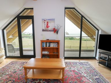 A living room with a wooden coffee table a patterned rug a bookcase and double glass doors overlooking a grassy area and the sea at Dunes in Rhosneigr