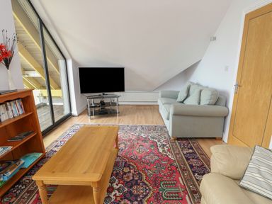 A living room with a wooden coffee table a patterned rug a gray sofa a beige armchair a bookshelf a flat screen tv and glass sliding doors at Dunes in Rhosneigr