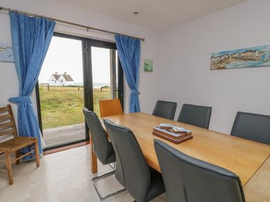 A dining room with a wooden table and chairs near glass doors with blue curtains showing a grassy outside view at Dunes in Rhosneigr