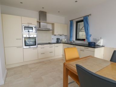 A kitchen with cream cabinets a built in oven and microwave a dining table with chairs and a window with blue curtains at Dunes in Rhosneigr