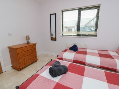 A bedroom with two single beds with red and white checkered bedding a wooden dresser with a lamp a wall mirror and a window with blinds at Dunes in Rhosneigr