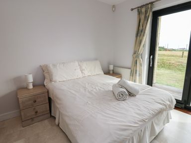 A bedroom with a bed with white bedding and rolled towels two bedside tables with lamps and a glass door with curtains at Dunes in Rhosneigr