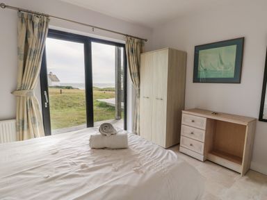 A bedroom with a bed towels wardrobe chest of drawers and a window with curtains showing a grassy outdoor area at Dunes in Rhosneigr