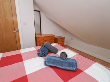 A bedroom with a bed covered by a red and white checkered blanket and rolled towels on top a wooden dresser and bedside table under a slanted ceiling