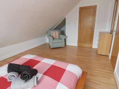 A bedroom with wooden floor a bed with red and white checkered blanket rolled towels an armchair near a door and a wooden dresser at Dunes in Rhosneigr