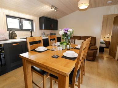 A dining area with a wooden table set with bowls glasses and flowers next to a kitchen with black cabinets at Enia Lodge 2 Llanelidan near Corwen
