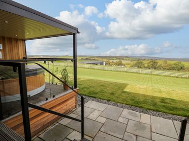 An outdoor patio with glass railing and a hot tub overlooking a grassy field and distant hills at Enia Lodge 2 in Llanelidan near Corwen