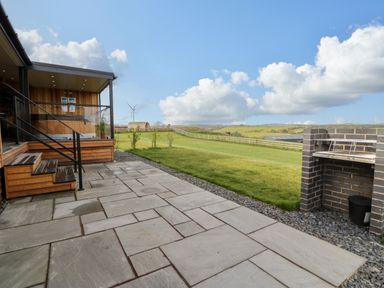 An outdoor patio with stone tiles and steps leading to a wooden structure with a hot tub and a brick barbecue grill on a lawn at Enia Lodge 2 in Llanelidan near Corwen