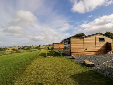 An outdoor scene with wooden lodges on gravel and grass with fields and hills in the background at Enia Lodge 2 Llanelidan near Corwen