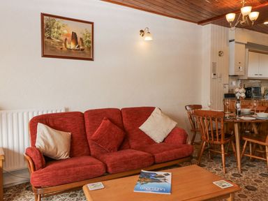 A living room with a red sofa and pillows a wooden coffee table a dining table with chairs set with plates and glasses at Lazey Cottage in Haverigg