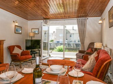 A dining table set with glasses and plates in a living room with armchairs and a television near a large window at Lazey Cottage in Haverigg