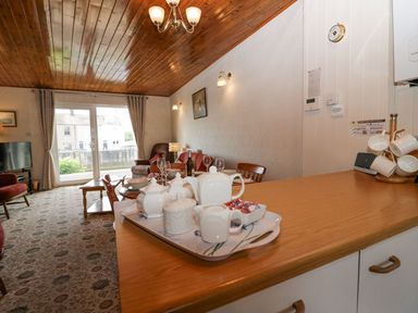 A living room with a wooden ceiling a coffee table chairs and a tray with teapots and cups on a kitchen counter at Lazey Cottage in Haverigg