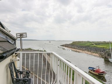 A balcony with a black metal chair overlooking a river with boats and a rocky shoreline at Lazey Cottage in Haverigg