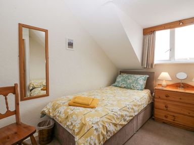 A small bedroom with a single bed with floral bedding a wooden chair a wooden chest of drawers with a lamp and mirror and a rectangular wall mirror at Lazey Cottage in Haverigg