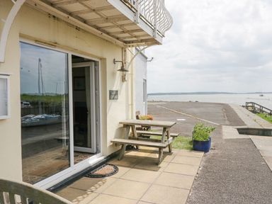 A patio area with a wooden picnic table outside a building by the water at Lazey Cottage in Haverigg