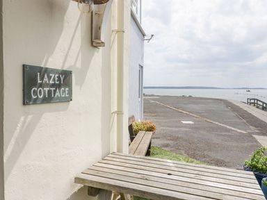 A wooden table and bench outside a building with a lazey cottage sign near a waterfront at Lazey Cottage in Haverigg
