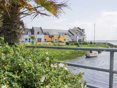 A waterfront view with a sailboat on the water colorful houses and greenery with a palm tree at Lazey Cottage in Haverigg