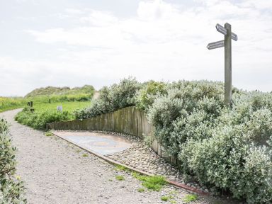A dirt path with a wooden signpost and green bushes alongside the path at Lazey Cottage in Haverigg