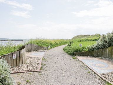 A gravel path between wooden fences and green shrubs by the water at Lazey Cottage in Haverigg