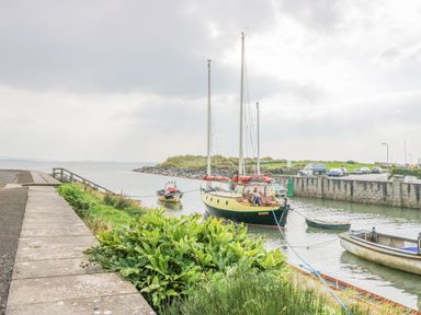 A harbor scene with sailboats and small boats tied at the dock by a walkway with plants and cars parked on the road at Lazey Cottage in Haverigg