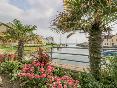 A waterfront view with palm trees and pink flowers along a railing near boats and houses at Lazey Cottage in Haverigg