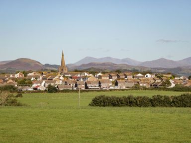 A village with houses and a church with a tall steeple surrounded by green fields and hills in the background at Lazey Cottage in Haverigg