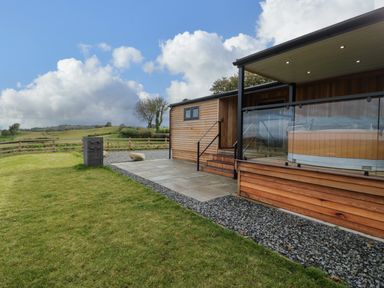 A wooden lodge with a covered porch and glass railing next to a grassy lawn and fenced field at Math Lodge 3 in Llanelidan near Corwen