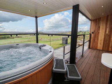 An outdoor covered patio with a hot tub wooden deck stairs glass railing and a grassy field beyond at Math Lodge 3 in Llanelidan near Corwen