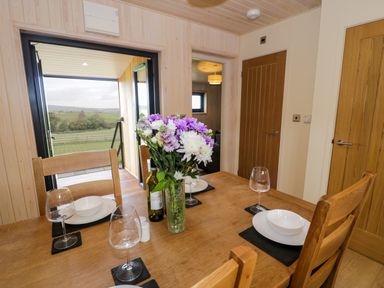 A dining table set with plates, bowls, wine glasses, a vase with flowers, and a bottle in a room with wooden walls and doors at Math Lodge 3 in Llanelidan near Corwen