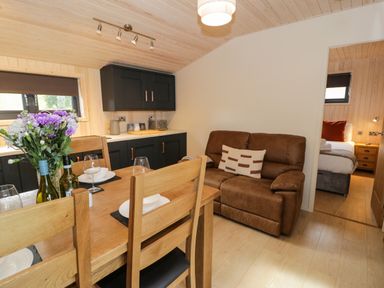 A dining table with flowers and wine bottles beside a brown sofa and a kitchen area at Math Lodge 3 in Llanelidan near Corwen