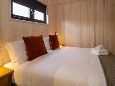 A bedroom with a bed featuring white sheets and red pillows beside a window with wooden walls at Math Lodge 3 in Llanelidan near Corwen