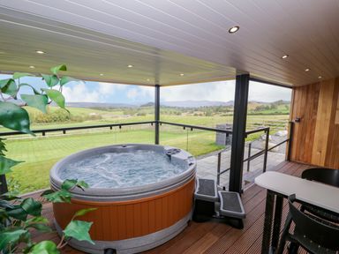An outdoor covered deck with a hot tub a table and chairs overlooking a grassy field and hills at Math Lodge 3 Llanelidan near Corwen