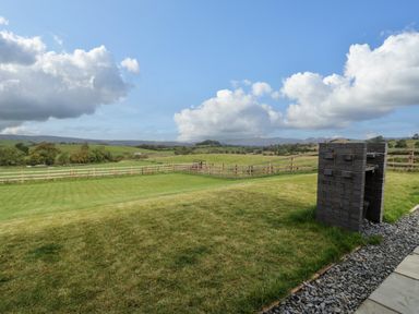 A grassy backyard with a brick barbecue and wooden fence with hills in the background at Math Lodge 3 in Llanelidan near Corwen