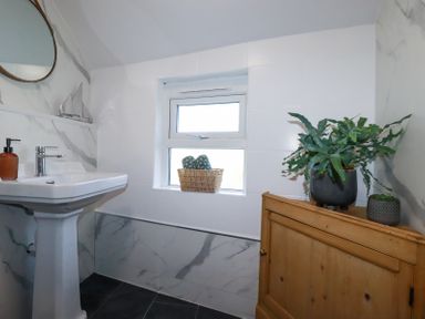 A bathroom with a pedestal sink a round mirror a window with a basket two towels on the sill and plants on a wooden cabinet at Hilldene Treknow