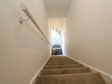 A carpeted staircase with a handrail on the left leading down to a wooden stool on a dark floor at Amelia's Point in St Ives