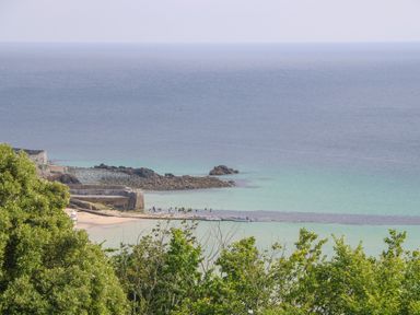 A coastal scene with green trees in the foreground a rocky shoreline and turquoise sea at Amelias Point in St Ives