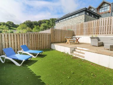 A fenced garden area with two blue sun loungers on grass and a raised patio with a table and outdoor seating at Ameilas Point in St Ives