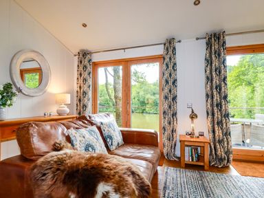 A living room with a leather sofa and patterned curtains by large glass doors overlooking a river at 10 Water's Edge in Lanreath