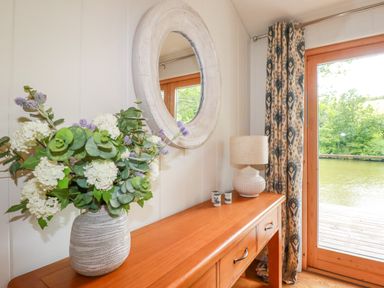A wooden table with a vase of flowers a lamp and a round wall mirror near glass doors with patterned curtains at 10 Water's Edge in Lanreath