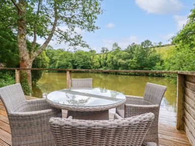 An outdoor deck with a glass top table and four wicker chairs overlooking a river with trees at 10 Water's Edge in Lanreath