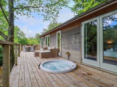 A wooden deck with a hot tub seating area and dining table surrounded by trees at 10 Water's Edge in Lanreath