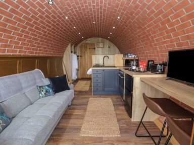 A narrow living space with a grey sofa and cushions a kitchen area with blue cabinets and a wooden countertop a wooden bar with two brown stools and a TV on the right Rhosydd Cyffylliog near Ruthin