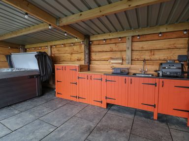 An outdoor kitchen with orange cabinets a sink a table-top grill and a hot tub under a covered wooden structure at Rhosydd in Cyffylliog near Ruthin