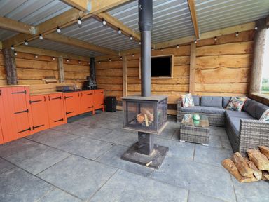 An outdoor covered seating area with a wood burning stove a grey wicker sofa set orange cabinets and string lights at Rhosydd in Cyffylliog near Ruthin