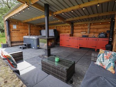 An outdoor covered lounge area with a hot tub a fireplace red cabinets a grill and sofas with cushions at Rhosydd in Cyffylliog near Ruthin