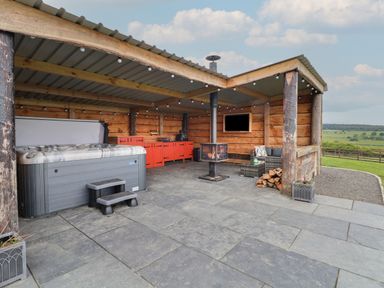 An outdoor covered patio area with a hot tub red cabinets a wood burning stove and seating with countryside views at Rhosydd in Cyffylliog near Ruthin