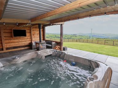 A covered outdoor space with a hot tub seating area wood stove and view of fenced green fields at Rhosydd in Cyffylliog near Ruthin