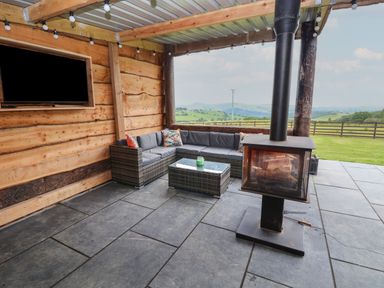 A covered outdoor seating area with a sectional sofa a glass table a wood stove and a mounted television with a view of green fields at Rhosydd in Cyffylliog near Ruthin