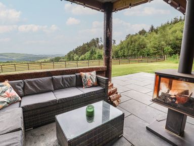 An outdoor seating area with a gray cushioned sofa and glass table near a wood-burning stove with a rural landscape at Rhosydd in Cyffylliog near Ruthin